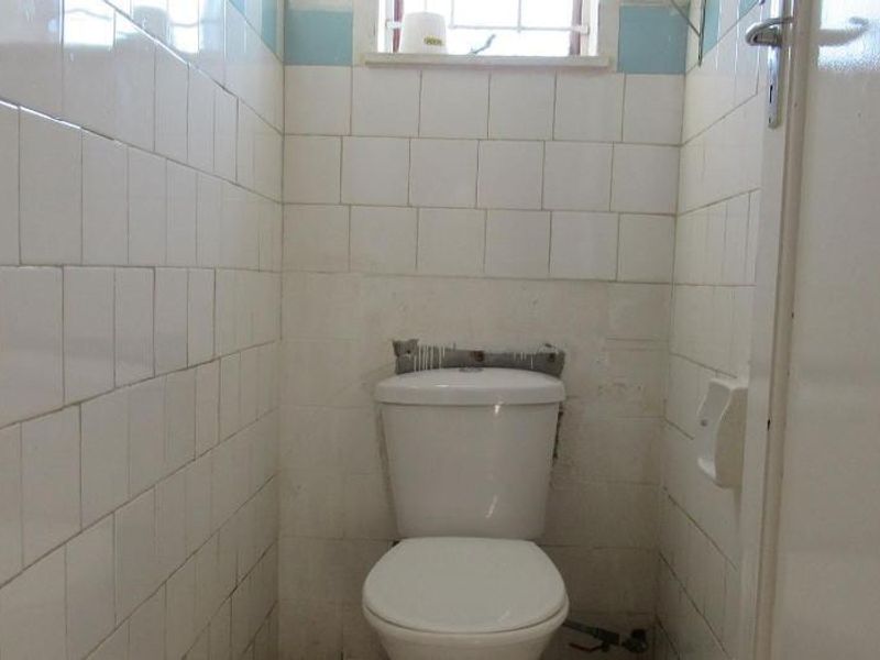 A small, dated bathroom featuring a toilet, tiled walls, and a window. The space requires renovation to address missing tiles and exposed pipework.