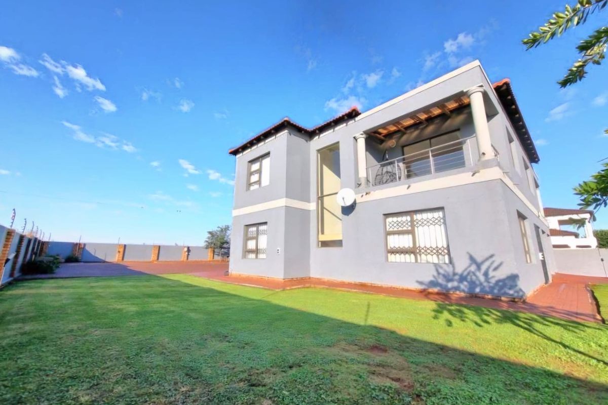 Rear view of a two-story house featuring a paved yard, lawn, balcony with columns, and secure perimeter fencing.
