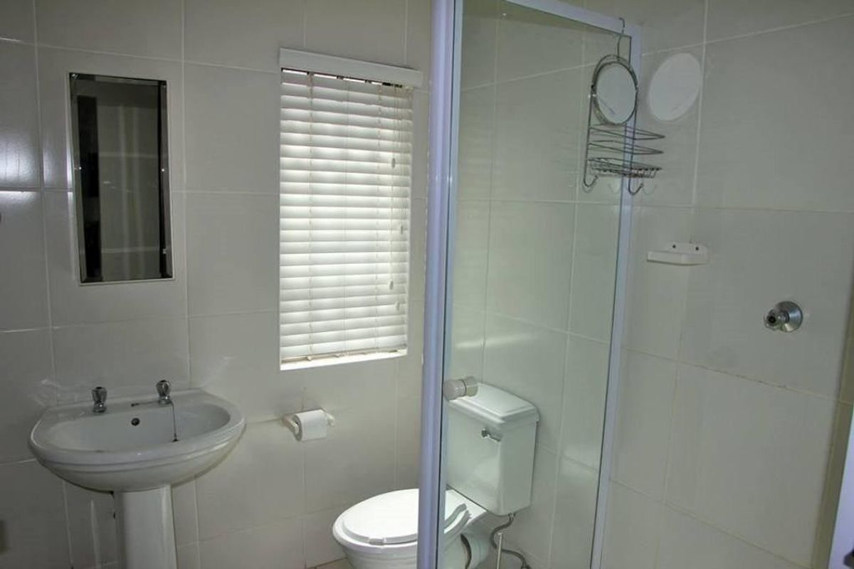 A functional bathroom featuring a pedestal basin, toilet, and a glass-enclosed shower with neutral wall tiling and a window with venetian blinds.