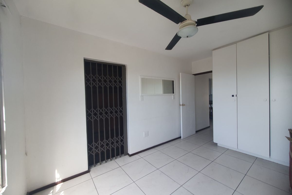 A bright, empty bedroom featuring tiled flooring, a ceiling fan, and built-in cupboards, with a security gate leading to an outdoor area.