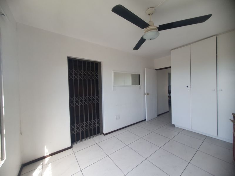 A bright, empty bedroom featuring tiled flooring, a ceiling fan, and built-in cupboards, with a security gate leading to an outdoor area.