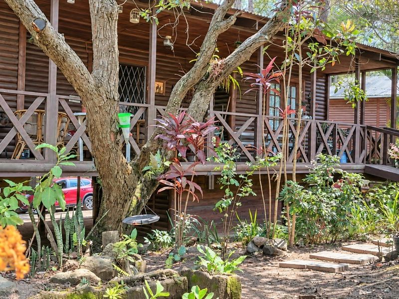 A rustic timber log townhouse featuring a raised wooden deck, balustrades, and glass doors, set within a natural garden environment.