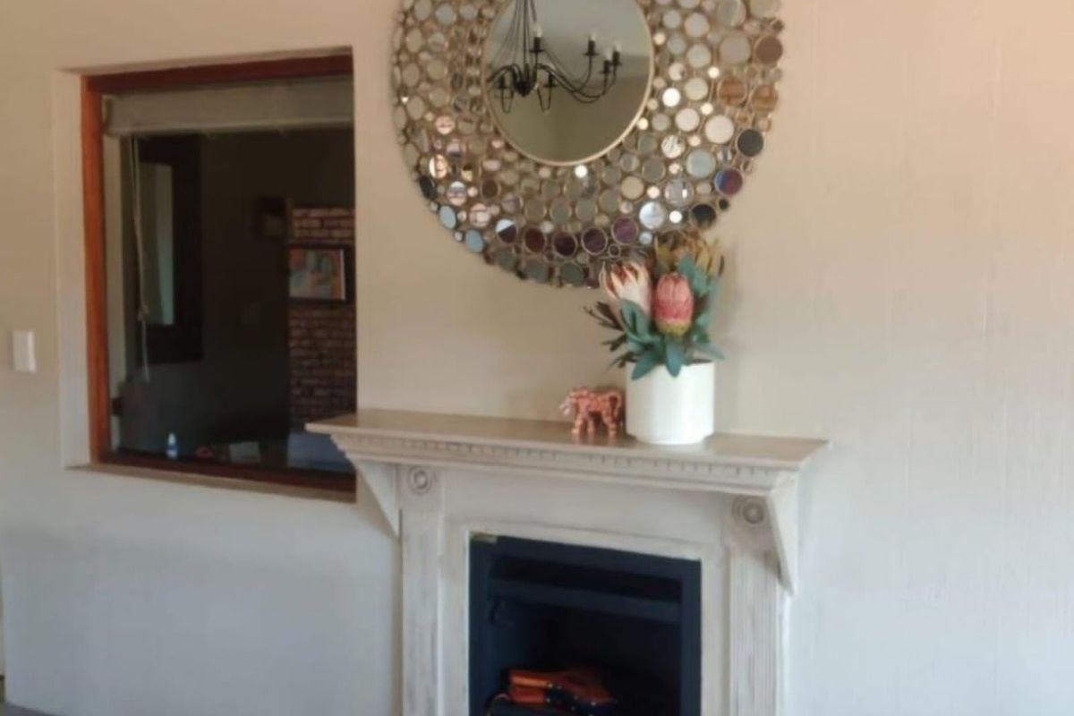 A living room featuring a decorative fireplace mantel and a large circular wall mirror, with a window looking into an adjacent space.