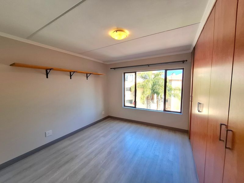 A bright, empty bedroom featuring light-toned laminate flooring, built-in wooden cupboards, and a wall-mounted shelf, offering a clean and neutral living space.