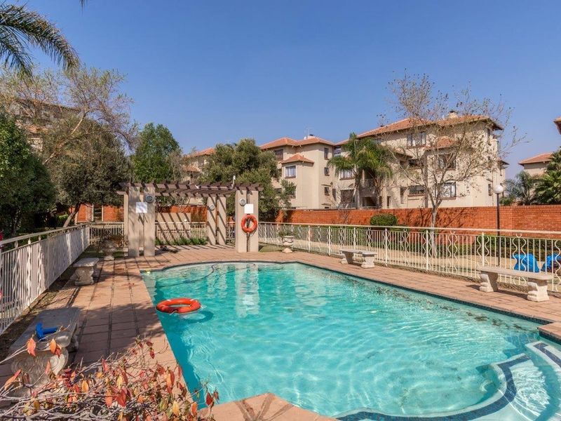 A well-maintained communal swimming pool area featuring a pergola, paved decking, and secure perimeter fencing within an apartment complex.