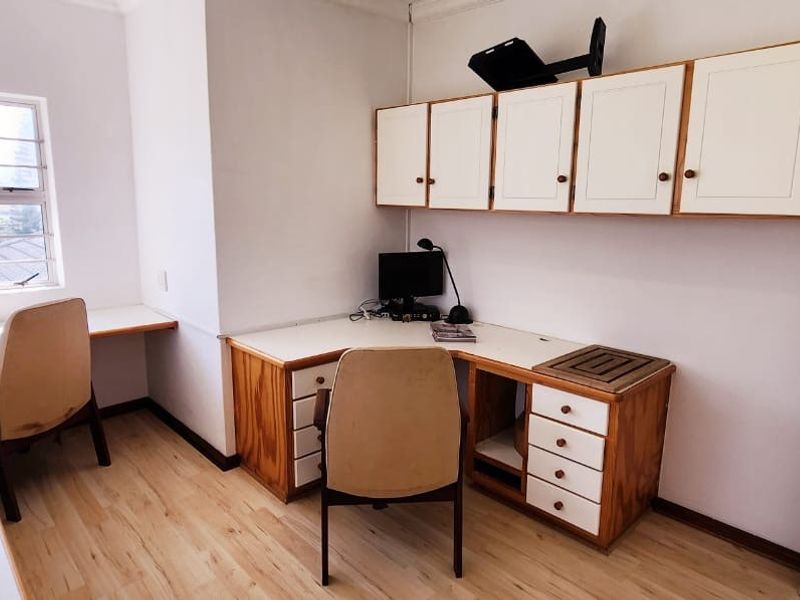 A functional home office featuring extensive built-in desk space, overhead storage cabinetry, and laminate flooring, providing a dedicated workspace with natural light.