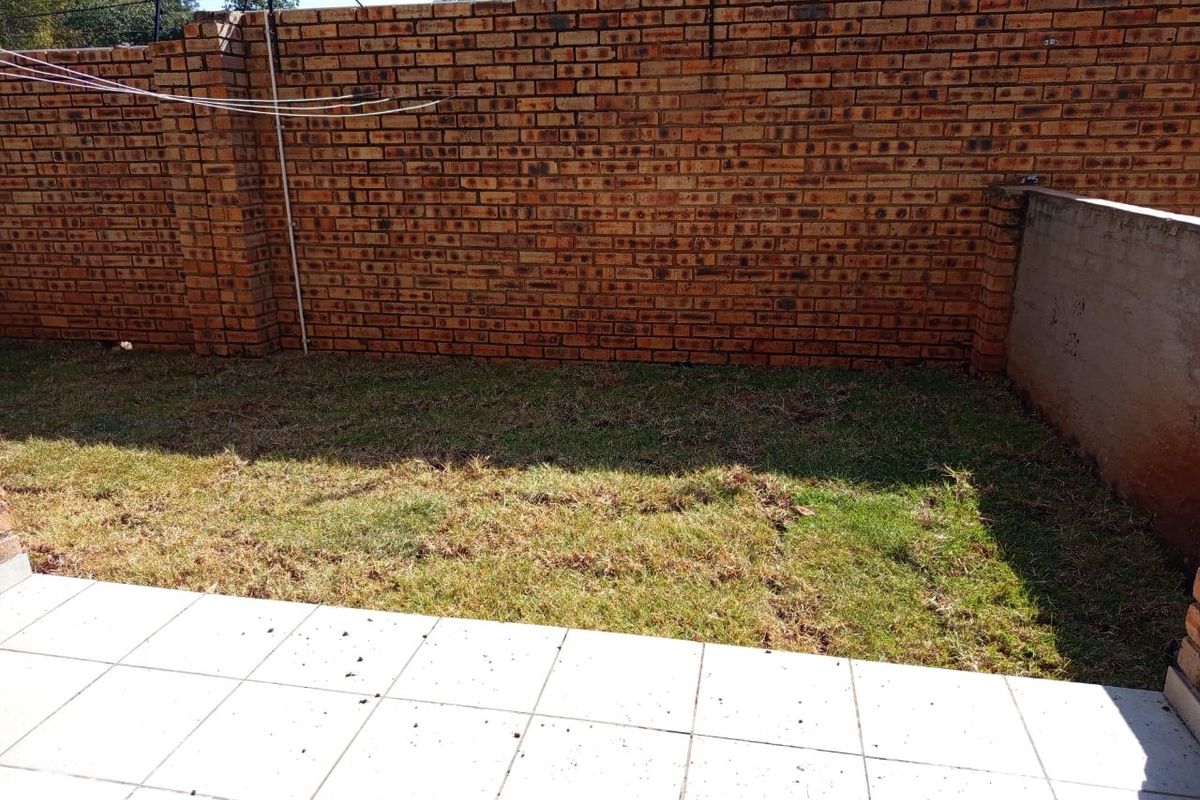 A private rear garden featuring a tiled patio area, a small lawn, and high brick boundary walls with security fencing.