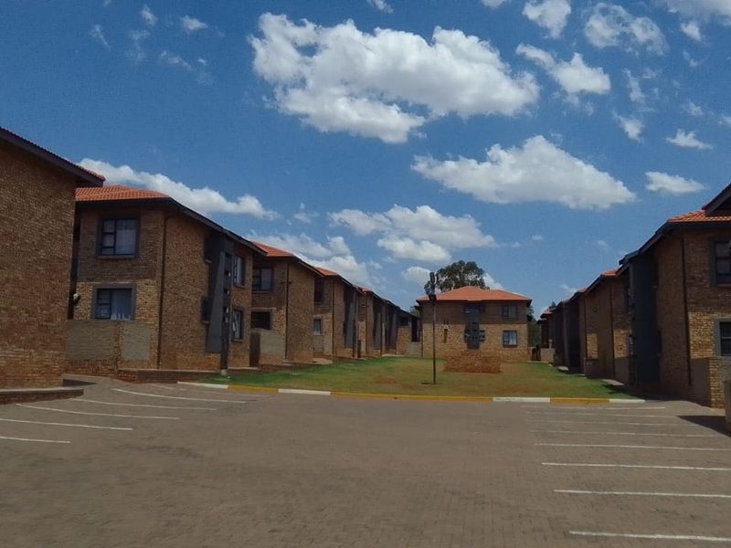 Exterior view of a residential apartment complex featuring face brick buildings, tiled roofs, and a paved parking area with communal garden space.