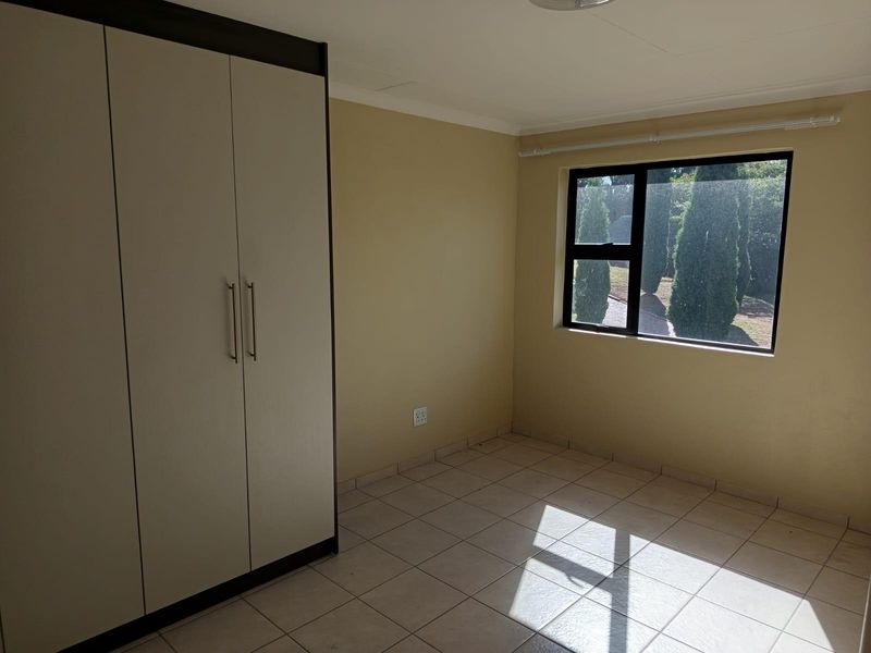 A bright, empty bedroom featuring neutral tiled flooring, a built-in wardrobe, and a large window providing natural light and garden views.