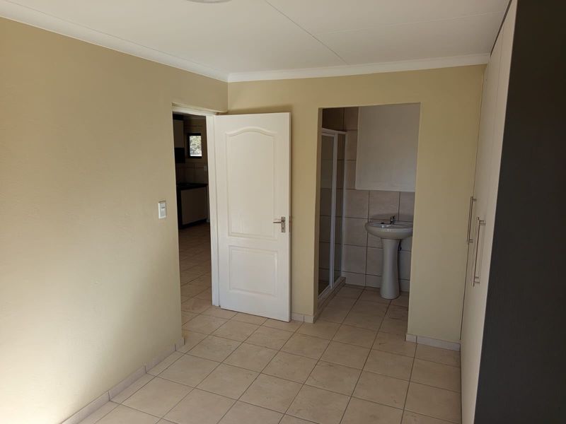 A spacious bedroom featuring neutral tiled flooring, built-in cupboards, and an en-suite bathroom equipped with a pedestal basin and shower cubicle.