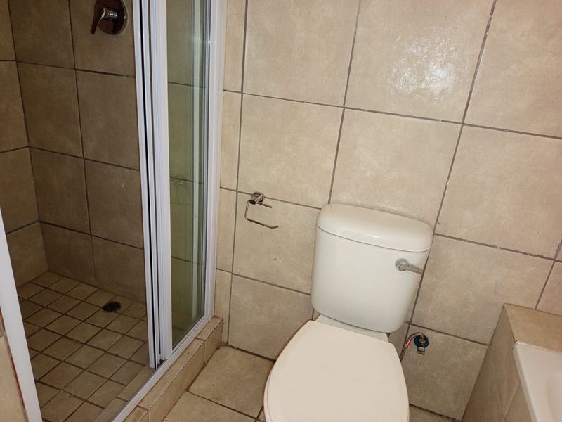 A functional bathroom featuring a shower enclosure and toilet with neutral ceramic tiling throughout, requiring some maintenance to the tile grout.