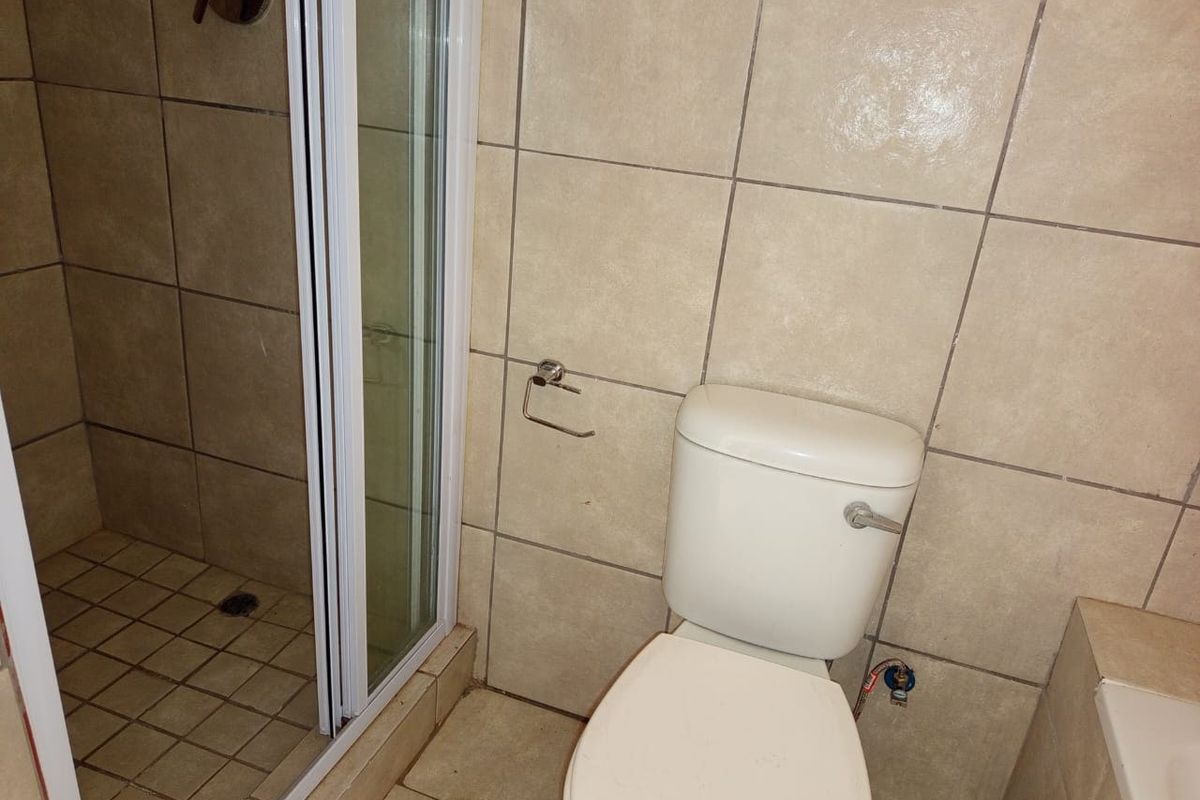 A functional bathroom featuring a shower enclosure and toilet with neutral ceramic tiling throughout, requiring some maintenance to the tile grout.