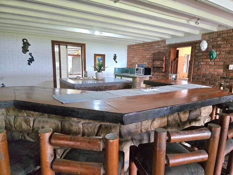 Rustic indoor bar area featuring a stone-clad counter, timber bar top, exposed brick walls, and an exposed beam ceiling with an integrated kitchenette area.