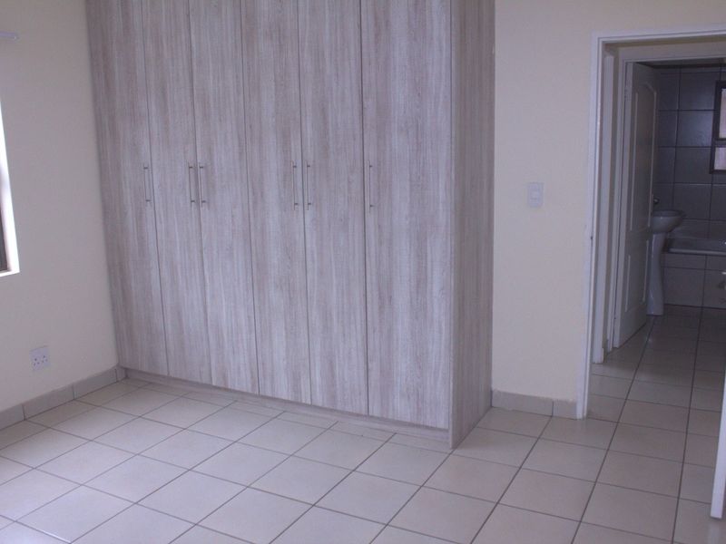 A clean, empty bedroom featuring light wood-look built-in cupboards, neutral tiled flooring, and direct access to an en-suite bathroom.