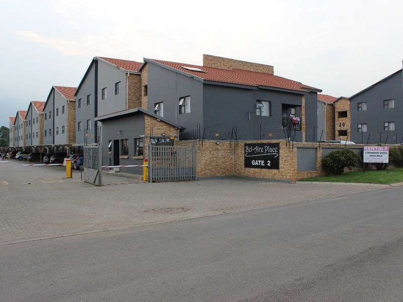 Exterior view of the Bel-Aire Place apartment complex entrance, featuring a paved driveway, security gate, and multi-story residential buildings with tiled roofs.