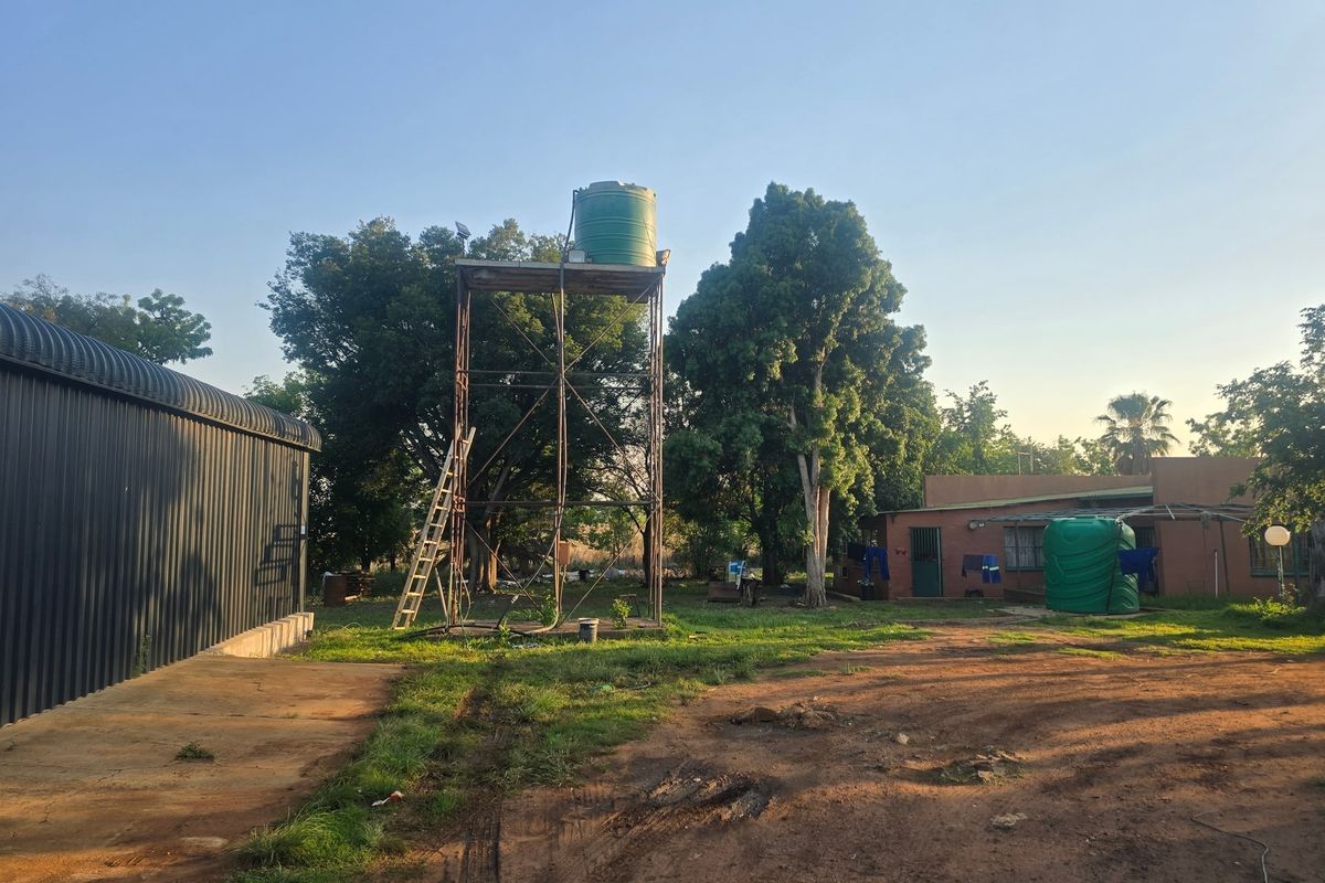 A farm yard view featuring a metal water tank stand, a corrugated metal shed, and a masonry dwelling set within a rural landscape.