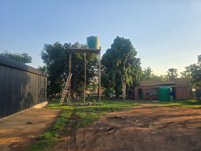 A farm yard view featuring a metal water tank stand, a corrugated metal shed, and a masonry dwelling set within a rural landscape.