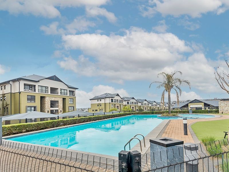 A well-maintained communal swimming pool area featuring paved decking, landscaped hedging, and perimeter fencing within a residential apartment complex.