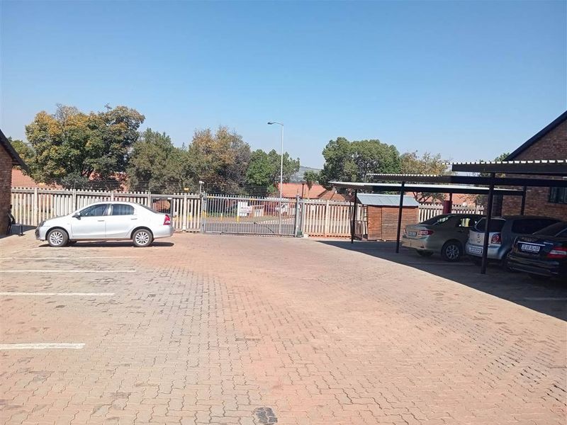 A paved communal parking area featuring a carport structure, secure perimeter fencing, and gated access to the townhouse complex.