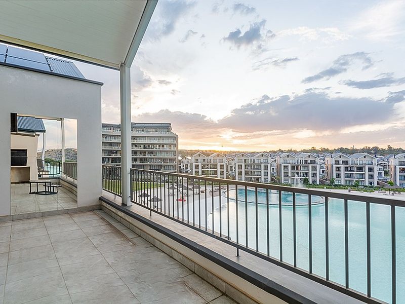 Spacious covered balcony featuring tiled flooring, a metal balustrade, and a built-in braai, offering scenic views of the estate pool and surrounding complex.