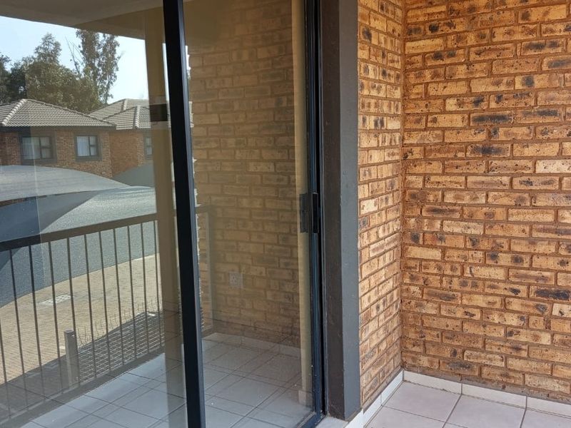 A private balcony featuring exposed brick walls, tiled flooring, and a sliding glass door entrance, offering a view of the complex parking area.
