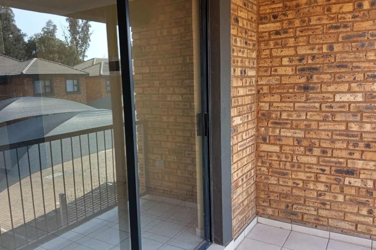A private balcony featuring exposed brick walls, tiled flooring, and a sliding glass door entrance, offering a view of the complex parking area.