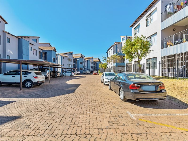 Exterior view of an apartment complex featuring paved parking areas, covered carports, and multi-story residential buildings with security fencing.