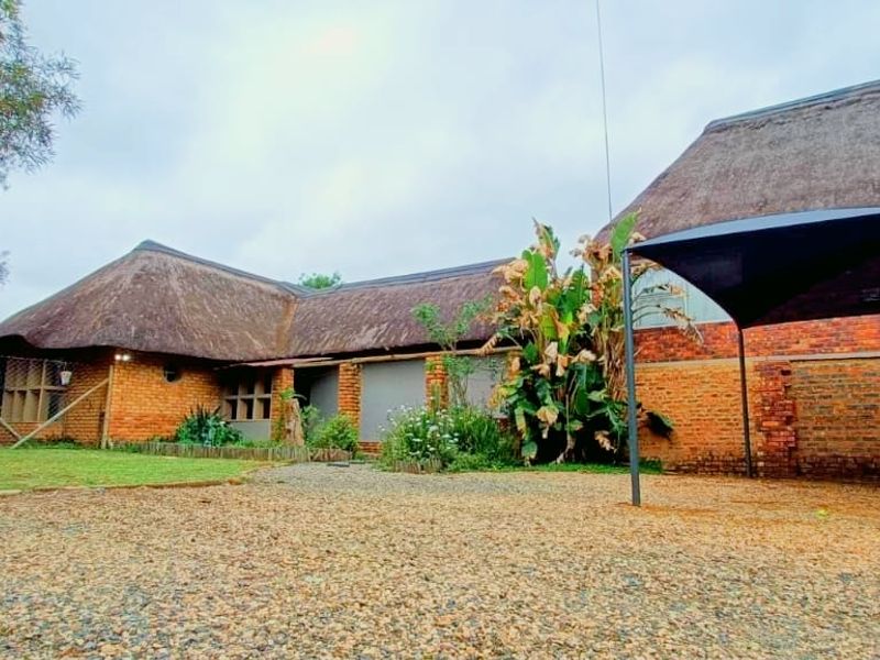 Exterior view of a rustic house featuring traditional thatch roofing, exposed brick facade, and a gravel driveway with a shade sail structure.