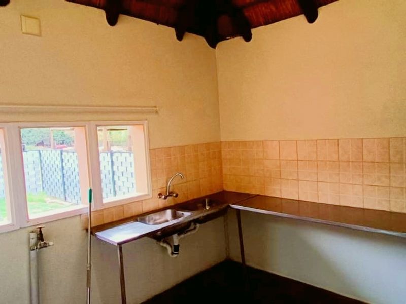 A functional scullery or utility area featuring a stainless steel sink, tiled splashback, and exposed metal work surfaces under a thatched roof.
