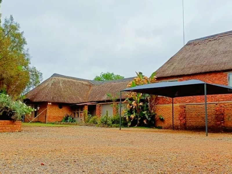 A charming brick house featuring traditional thatched roofing, a gravel driveway, and a covered carport area set within a landscaped garden.
