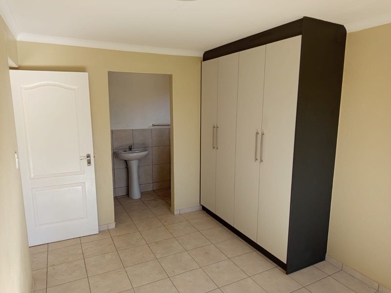 A clean, empty bedroom featuring tiled flooring, a large built-in wardrobe, and an adjoining en-suite area with a pedestal basin.