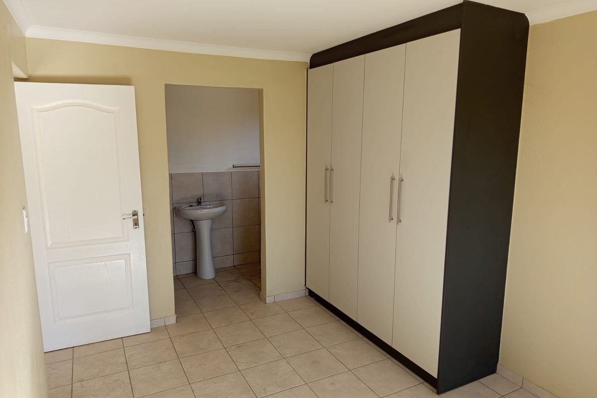 A clean, empty bedroom featuring tiled flooring, a large built-in wardrobe, and an adjoining en-suite area with a pedestal basin.