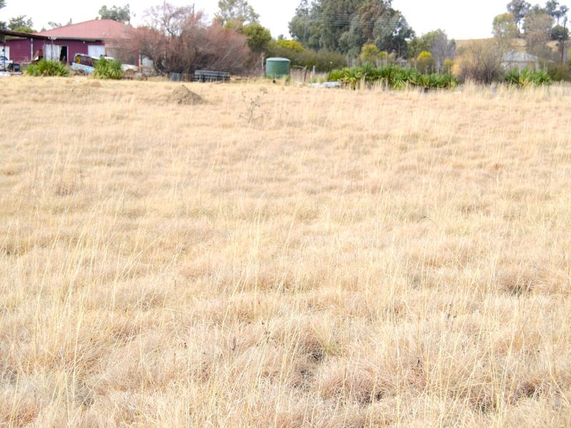 A wide view of a vacant residential plot featuring dry grass, a water tank, and distant outbuildings.