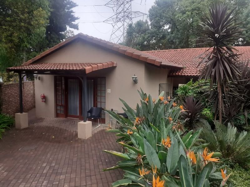 A rear view of the apartment featuring a covered patio area with brick paving, timber-framed glass doors, and established garden landscaping.