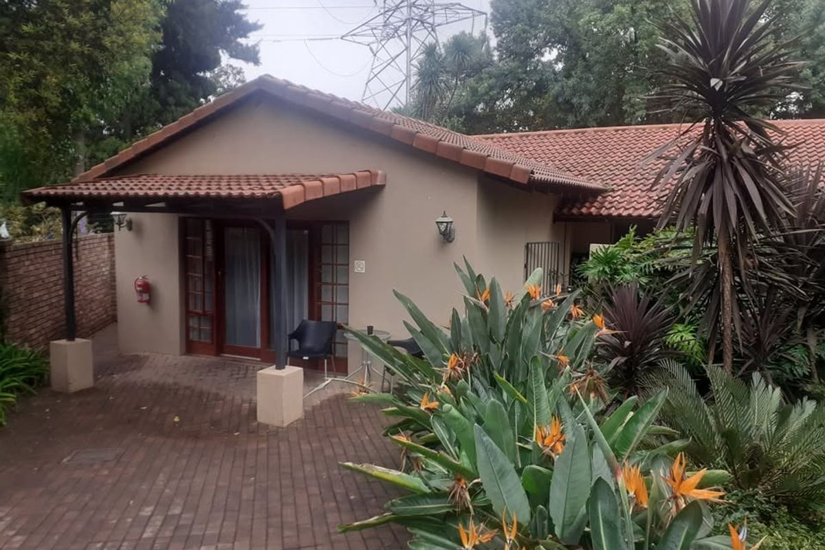 A rear view of the apartment featuring a covered patio area with brick paving, timber-framed glass doors, and established garden landscaping.