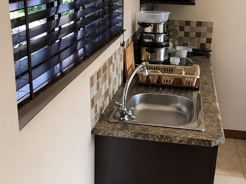 A compact scullery or prep area featuring a stainless steel sink, laminate countertop, tiled splashback, and venetian blinds.