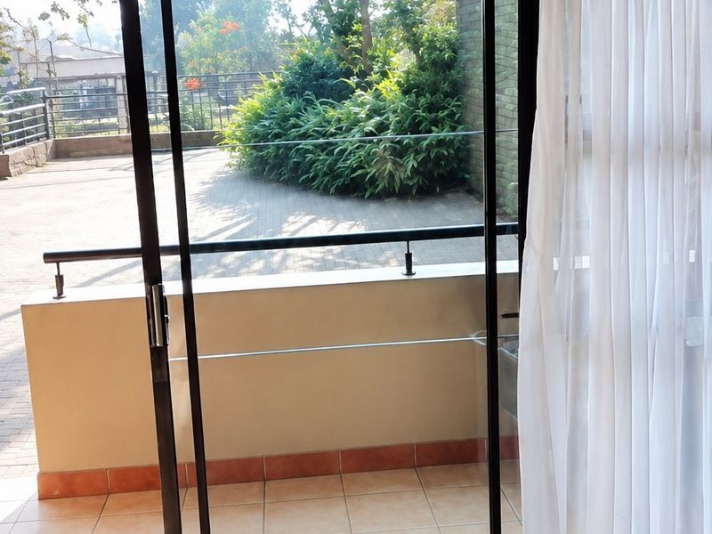 A view from an interior space through a sliding glass door onto a tiled balcony with a metal balustrade and garden outlook.