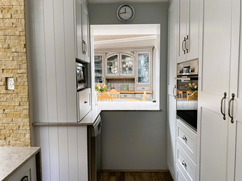 A well-appointed scullery featuring custom cabinetry, stone countertops, and integrated appliances, with a convenient serving hatch looking through to the dining area.