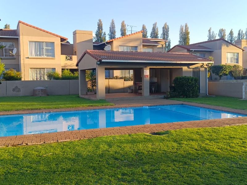A well-maintained communal pool area featuring a paved surround, manicured lawn, and a covered patio structure within an apartment complex setting.