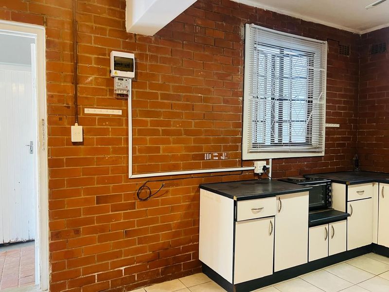 A kitchen area featuring exposed brick walls, fitted cabinetry with laminate countertops, and tiled flooring, requiring cosmetic updates to improve the aesthetic appeal.