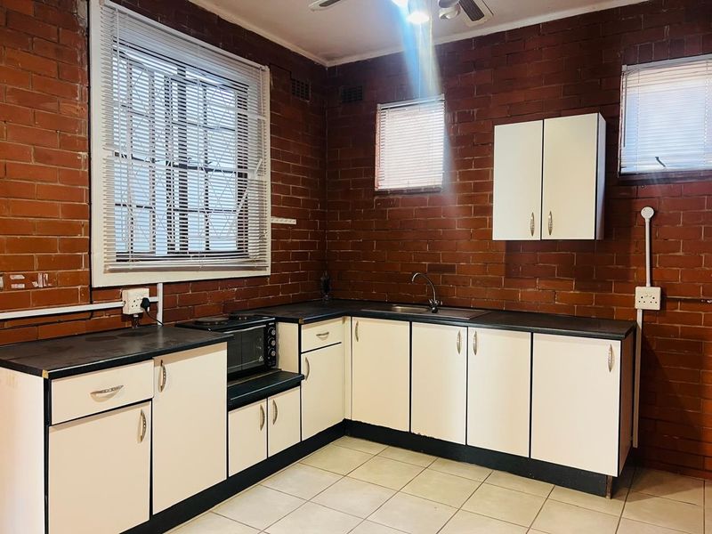 A functional kitchen featuring exposed brick walls, white cabinetry, dark laminate countertops, and tiled flooring with surface-mounted electrical fixtures.