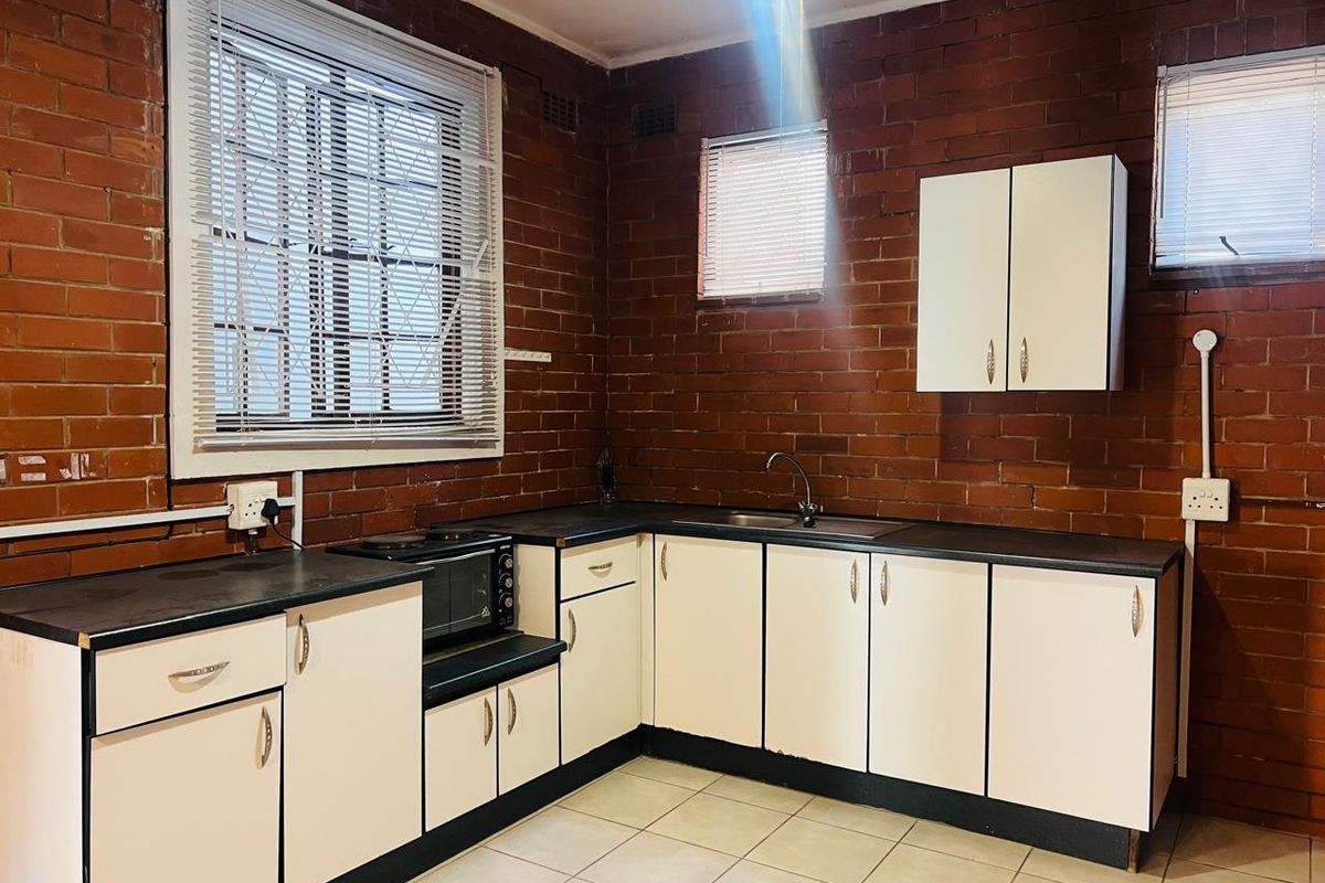 A functional kitchen featuring exposed brick walls, white cabinetry, dark laminate countertops, and tiled flooring with surface-mounted electrical fixtures.