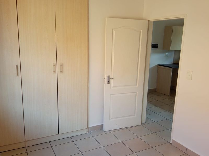 A clean, empty bedroom featuring light wood-finish built-in cupboards, tiled flooring, and direct access to an adjacent kitchen area.