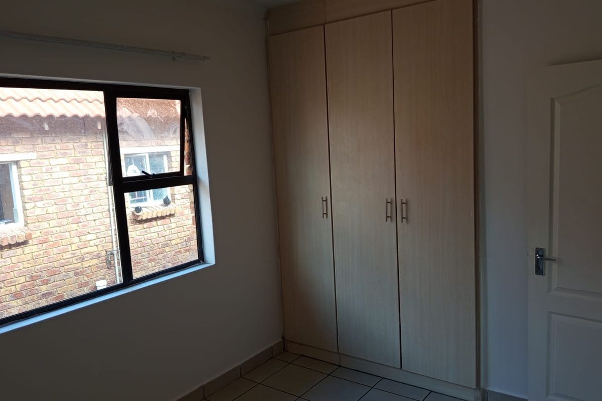A clean, empty bedroom featuring built-in cupboards, tiled flooring, and an aluminium-framed window overlooking the exterior.