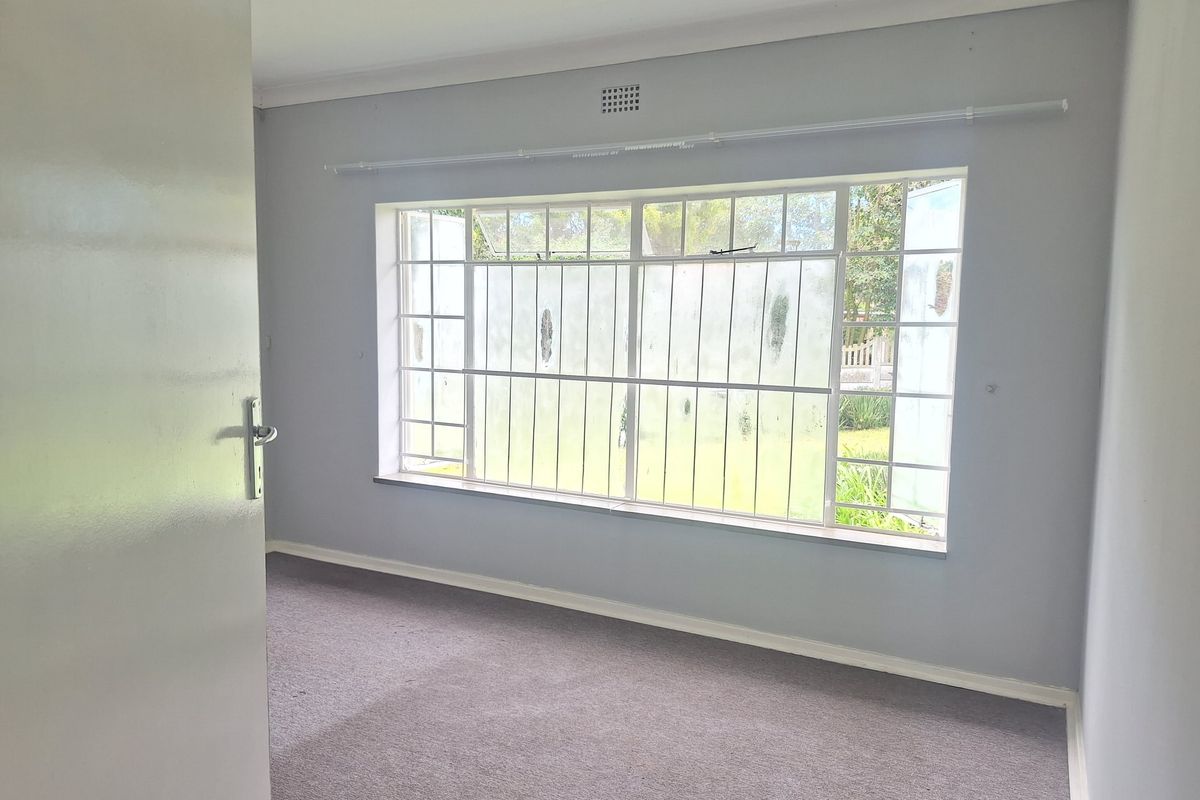 A bright, empty bedroom featuring neutral grey walls, carpeted flooring, and a large window fitted with security bars and a curtain rail.