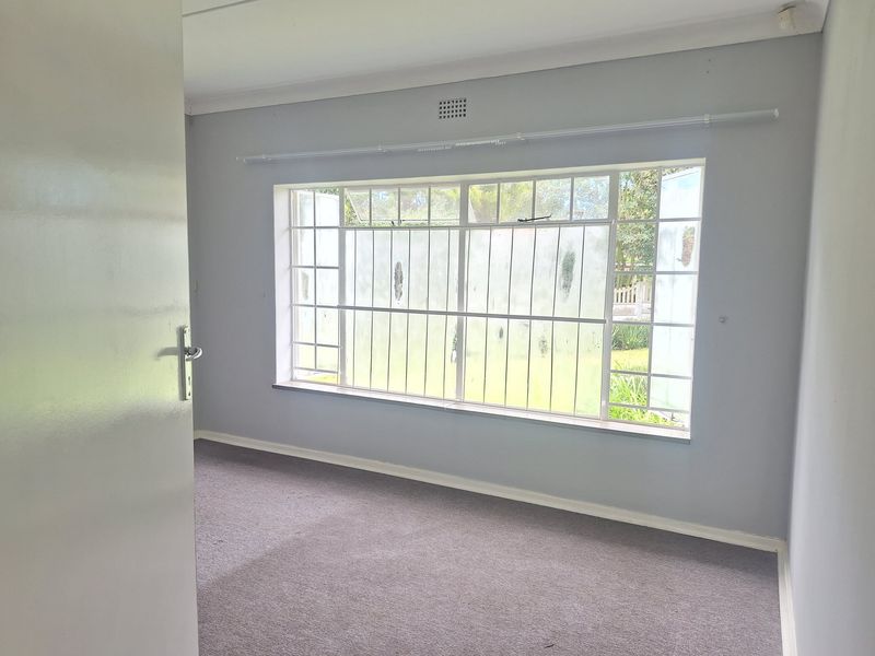 A bright, empty bedroom featuring neutral grey walls, carpeted flooring, and a large window fitted with security bars and a curtain rail.
