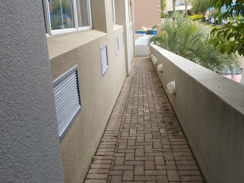 A narrow paved exterior walkway along the side of the commercial building, featuring wall-mounted lighting and ventilation grilles.