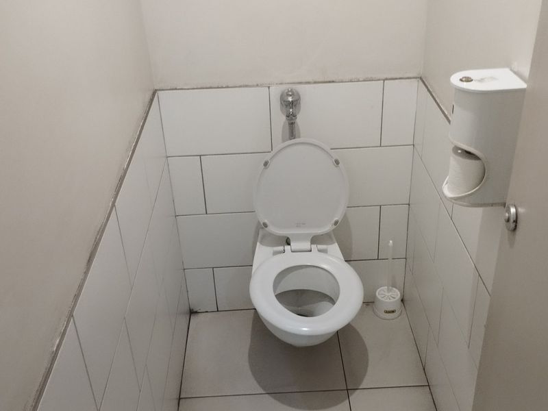 A compact commercial restroom featuring white wall and floor tiling, a standard toilet, and a wall-mounted toilet paper dispenser.