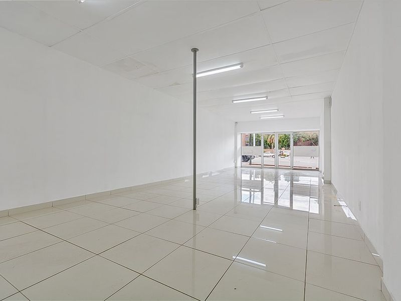 A bright, open-plan commercial space featuring polished tiled flooring, a suspended ceiling with fluorescent lighting, and a large glass storefront entrance.