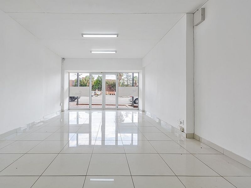 A bright, empty commercial office space featuring polished white floor tiles, fluorescent lighting, and a glass entrance door facing the street.